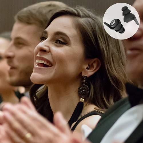 Lächelnde Frau in elegantem Outfit trägt diskrete schwarze Ohrstöpsel während eines Konzerts. Die Ohrstöpsel sind besonders geeignet für Musikgenuss und bieten angenehmen Gehörschutz, ohne die Klangqualität zu beeinträchtigen.